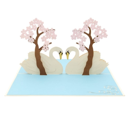 This 3-D pop-up card shows a swan couple in love, their necks curved together, forming a heart shape. With pink flowering trees and a baby blue lake, this card shimmers with affection.
